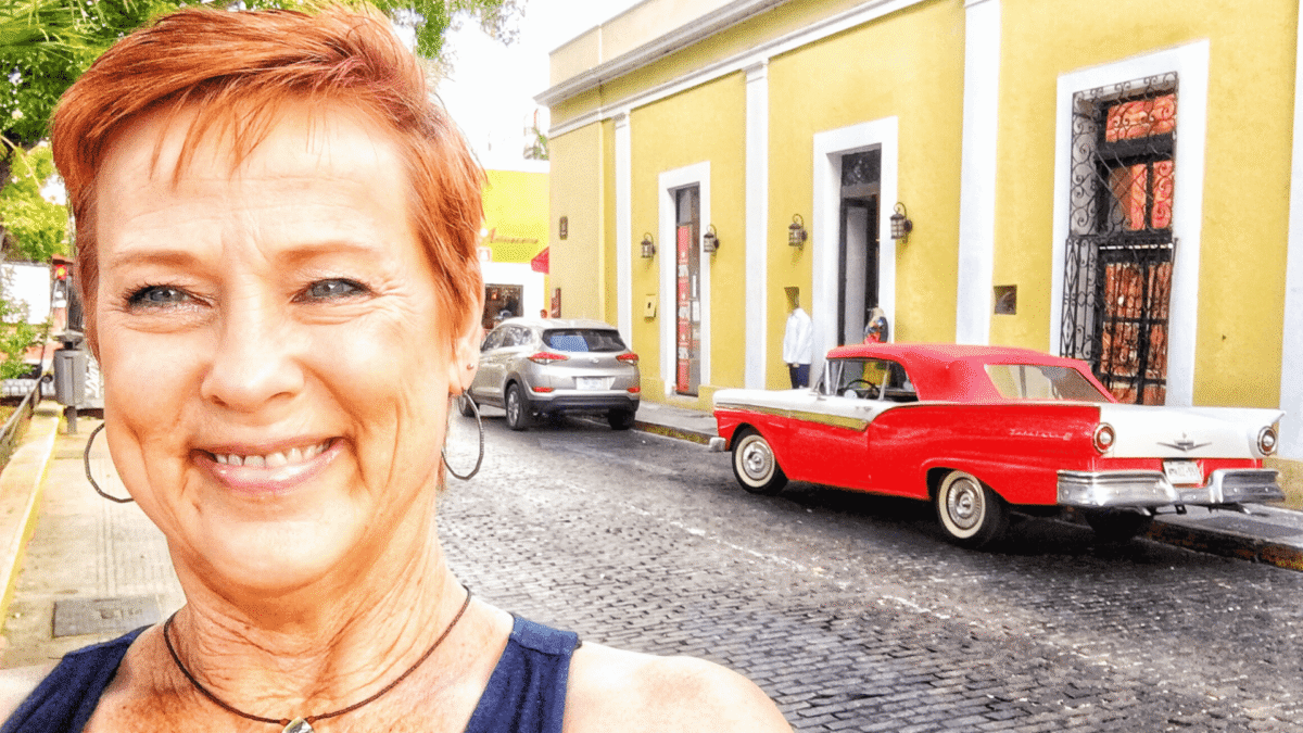 A smiling selfie of Amy on a historic cobblestone street in Merida, Mexico, featuring a vintage red car and bright yellow colonial buildings.