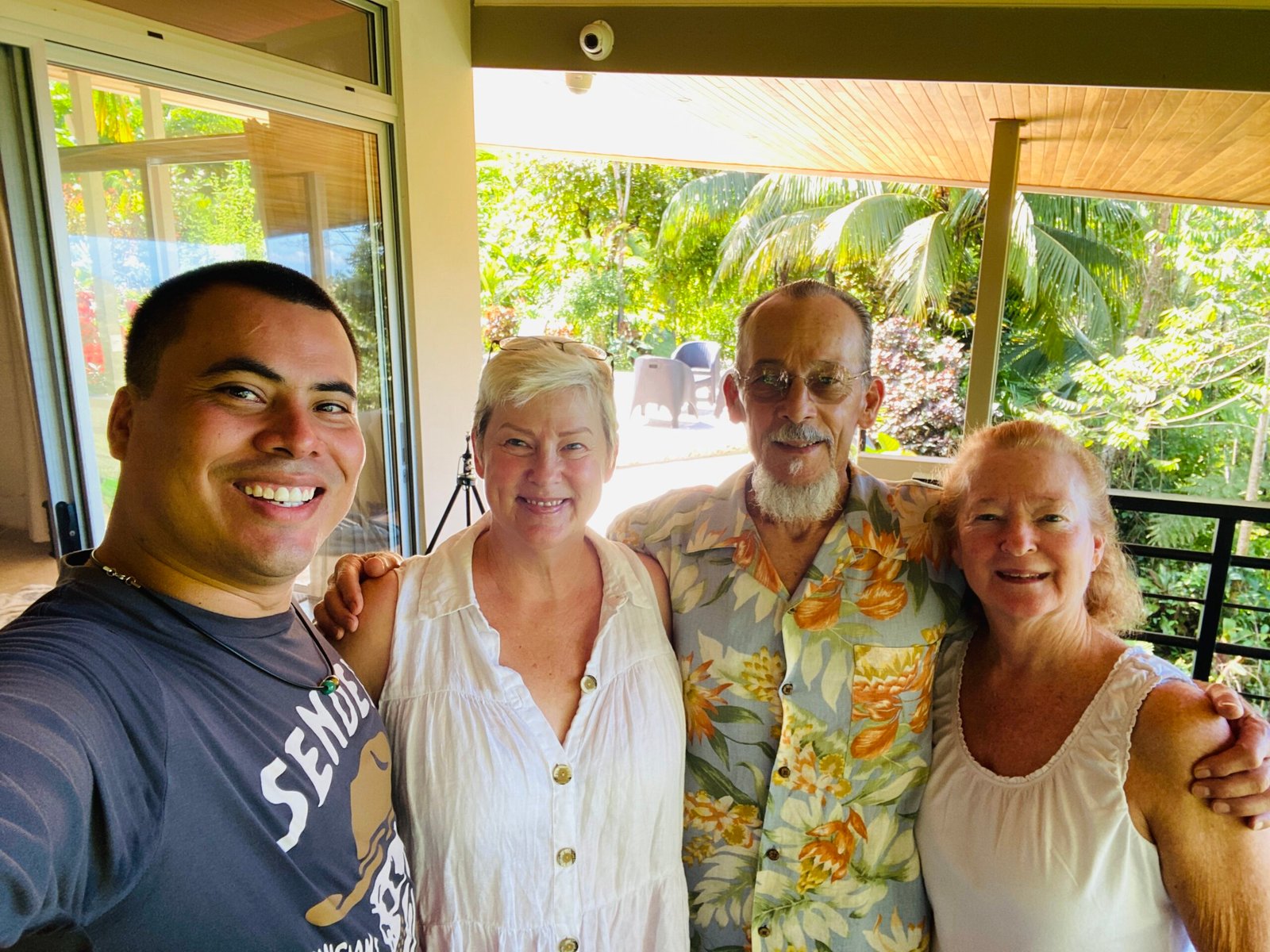 A smiling group photo of Amy, Angel, and friends embracing on a shady outdoor patio in Merida surrounded by lush green palm trees.