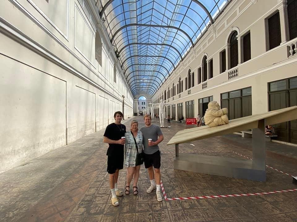 Amy and two young men standing in a modern, brightly lit shopping arcade in Merida, Mexico, which features a large glass skylight ceiling.