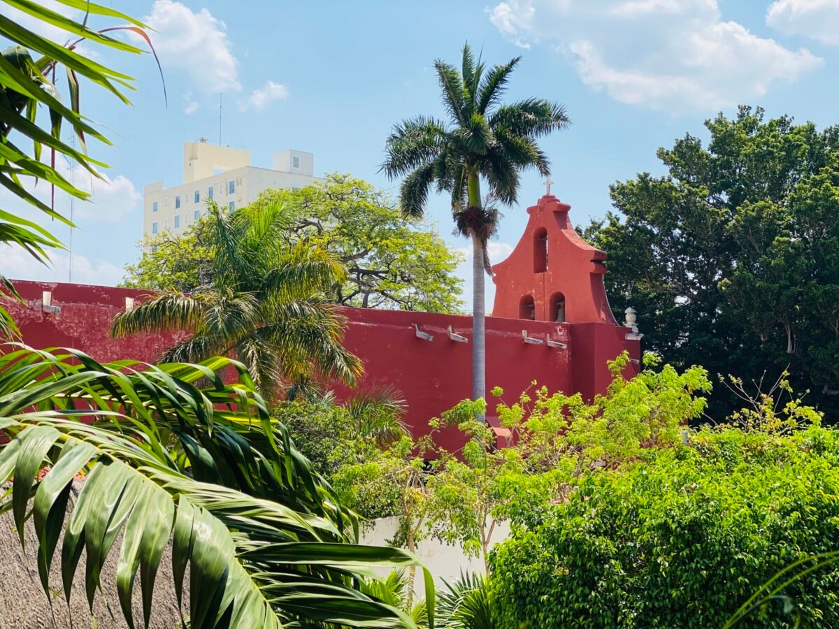 Red colonial church surrounded by tropical palms and lush greenery in Mérida, Mexico
