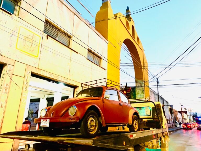 Red volkswagon beetle being towed underneath a yellow arch in Merida Mexico centro